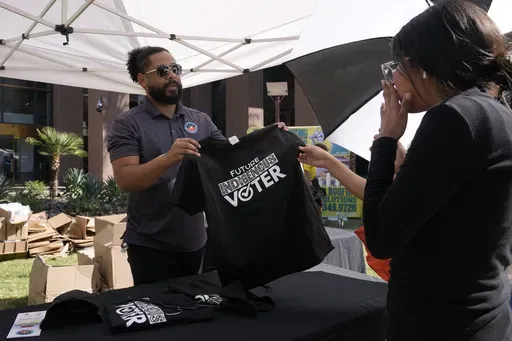 Alexander Castillo-Nunez, left, a civic engagement coordinator at the Inter Tribal Council of Arizona, Inc., shows a voter t-shirt at an Arizona Native Vote booth during an Indigenous Peoples' Day event Monday, Oct. 14, 2024, in Phoenix. (AP Photo/Ross D. Franklin)