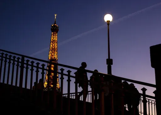 People walk on a bridge next to the Eiffel Tower in Paris, Wednesday Feb. 9, 2022. Lights on the Eiffel Tower will soon be turned off an hour earlier at night as part of an energy savings plan in the French capital, its mayor announced. Paris mayor said the iconic tower that is illuminated until 1:00am is only one of the city's monuments and municipal buildings that will be plunged into darkness earlier in the evening as the French capital faces risks of power shortages, rationing and blackouts 