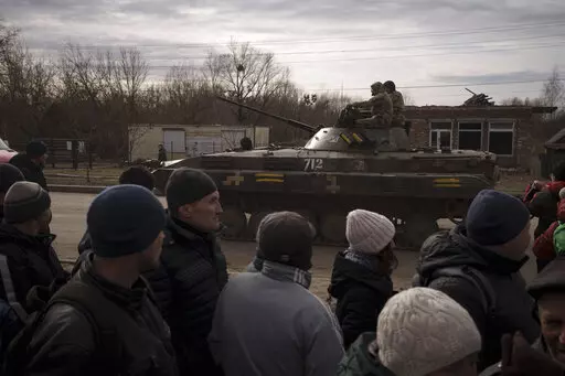 Residents lining up for aid watch as Ukrainian soldiers ride atop a tank in the town of Trostsyanets, Ukraine, Monday, March 28, 2022. Trostsyanets was recently retaken by Ukrainian forces after being held by Russians since the early days of the war. (AP Photo/Felipe Dana)