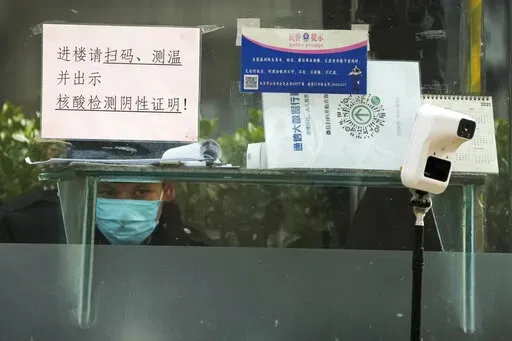 A security guard keeps watch inside a booth displaying a temperature scan device and a travel QR code, outside an office building in Beijing, Monday, Dec. 12, 2022. China will drop a travel tracing requirement as part of an uncertain exit from its strict "zero-COVID" policies that have elicited widespread dissatisfaction. (AP Photo/Andy Wong)