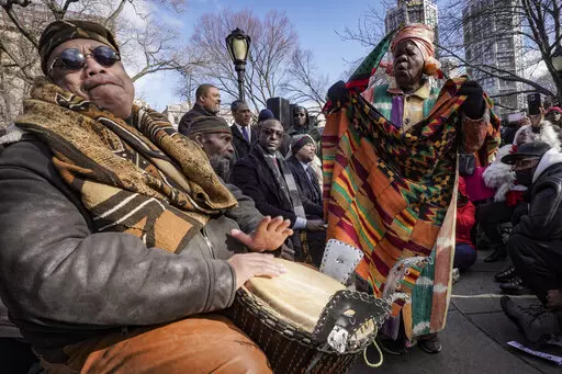 Master drummer Baba Don Babatunde, far left, and civil rights activist Queen Mother Moore, far right, lead a spiritual moment, during the Central Park gate-naming ceremony to honor the five men exonerated after being wrongfully convicted as teenagers for the 1989 rape of a jogger in Central Park, including Yusef Salaam, seated third from left, Raymond Santana Jr., seated fourth from left, Monday Dec. 19, 2022, in New York. (AP Photo/Bebeto Matthews)