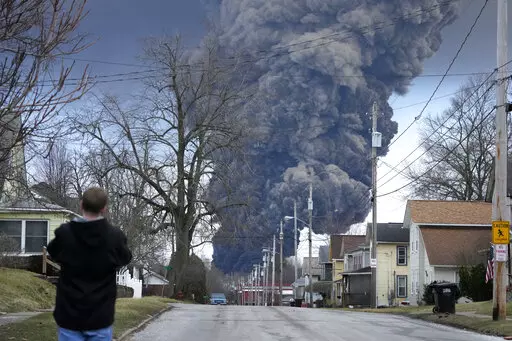 A man takes photos as a black plume rises over East Palestine, Ohio, as a result of a controlled detonation of a portion of the derailed Norfolk Southern train, Feb. 6, 2023. After toxic chemicals were released into the air from a wrecked train in Ohio, evacuated residents remain in the dark about what toxic substances are lingering in their vacated neighborhoods while they await approval to return home. (AP Photo/Gene J. Puskar, File)