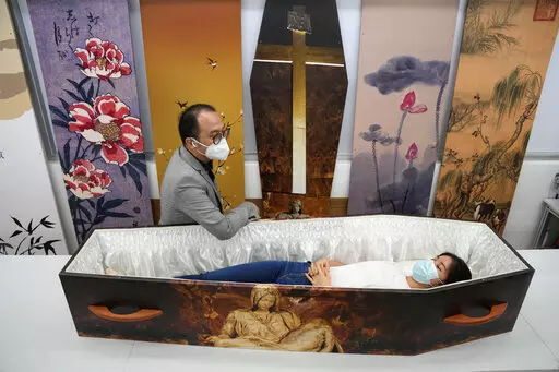 Wilson Tong, CEO of LifeArt Asia, left, talks to a reporter who tries out a catholic-designed paper coffin at Tong's factory in Hong Kong, Friday, March 18, 2022. Hong Kong is running short of coffins during its deadliest outbreak of the coronavirus pandemic, which has cost about 6,000 lives so far this year.  LifeArt, a company in Hong Kong is trying to make an alternative, cardboard coffin, which it says is environmentally-friendly. (AP Photo/Kin Cheung)