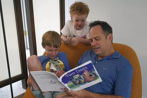 Wes Brown, right, reads to children Shawn Larimer-Brown, 7, left, and Charlie Larimer-Brown, 5, center, at their home Friday, Aug. 18, 2023, in Winter Park, Fla. Across the country, books and lessons that represent different families and identities to the youngest of learners are increasingly the target of the conservative pushback to efforts to promote diversity and inclusion. (AP Photo/John Raoux)