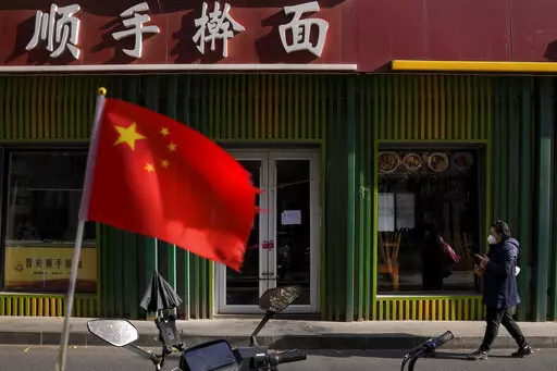 A woman wearing a face mask walks past a national flag outside a closed noodles restaurant due to the COVID-19 controls in Beijing, Wednesday, Nov. 23, 2022. (AP Photo/Andy Wong, File)