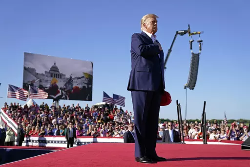 As footage from the Jan. 6, 2021, insurrection at the U.S. Capitol is displayed in the background, former President Donald Trump stands while a song, "Justice for All," is played during a campaign rally at Waco Regional Airport, Saturday, March 25, 2023, in Waco, Texas. The song features a choir of men imprisoned for their role in the Jan. 6, 2021, insurrection singing the national anthem and a recording of Trump reciting the Pledge of Allegiance. (AP Photo/Evan Vucci)