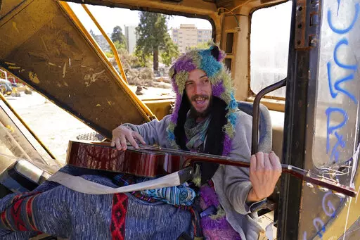 At Atmaa Das, 28, who is homeless from Alabama, strums his guitar while singing parts of Woody Guthrie's anthem, "This Land Is Your Land" while sitting in a piece of construction equipment left behind at People's Park in Berkeley, Calif., on Aug. 16, 2022. The three-acre site's colorful history, forged from University of California, Berkeley's seizure of the land in 1968, has been thrust back into the spotlight by the school's renewed effort to pave over People's Park as part of a $312 million p