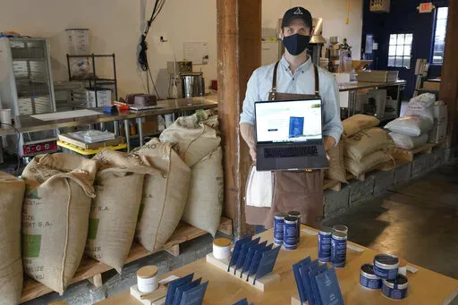 Kelly Van Arsdale poses for a photo Monday, Jan. 31, 2022, in Spinnaker Chocolate, the store he owns with his brother in Seattle where they make and sell single-origin chocolate bars. Van Arsdale is holding a laptop showing his company's website, and he says despite getting a lot of foot-traffic business, reaching customers online has been expensive and difficult. (AP Photo/Ted S. Warren)