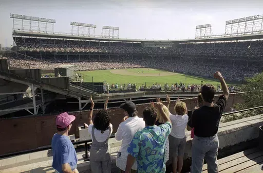 In this Monday, July 9, 1990, file photo, spectators watch an All-Star Game practice session from the roof of a building just outside Chicago's Wrigley Field. Booking hotels for baseball games during shoulder seasons like May, June or September can often be cheaper than the summer months. Additionally, save money by planning to attend weekday versus weekend games. (AP Photo/Seth Perlman, File)