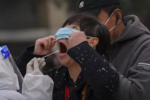A woman pulls up her mask to get her throat swab at a coronavirus testing site near  residential buildings, Wednesday, April 6, 2022, in Beijing. (AP Photo/Andy Wong)