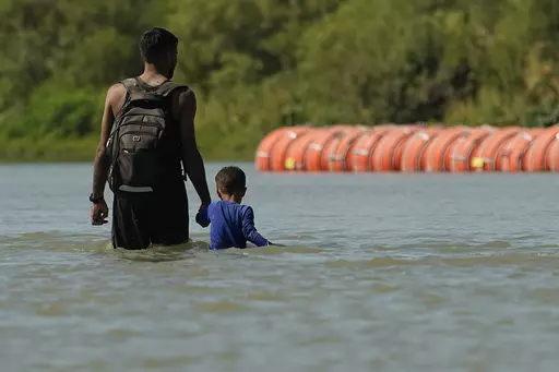 Migrants walk past large buoys being used as a floating border barrier on the Rio Grande, Aug. 1, 2023, in Eagle Pass, Texas. Texas attorneys asked federal appeals court judges Thursday, Oct. 5, 2023 to let the state keep in place a floating barrier of large concrete-anchored buoys to block migrants from crossing the Rio Grande — a barrier the Biden administration says was illegally deployed without needed federal authorization. (AP Photo/Eric Gay, file)