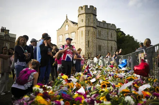People gather at flowers and messages to tribute Queen Elizabeth II, in front of Windsor Castle in Windsor, England, Sunday, Sept. 11, 2022. Queen Elizabeth II, Britain's longest-reigning monarch and a rock of stability across much of a turbulent century, died Thursday Sept. 8, 2022, after 70 years on the throne. She was 96. (AP Photo/Markus Schreiber)