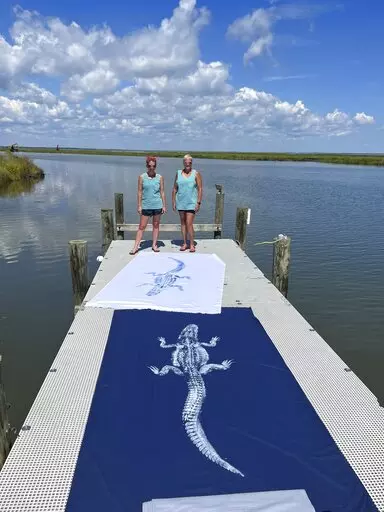 Robin Miller Leslie Charleville, left, stands next to her business partner Cindy Verdin while displaying two gyokatu alligator prints made at alligator hunter Logan Davis' dock near The Rigolets on August, 31, 2022, outside Slidell, La. (Robin Miller/The Advocate via AP)